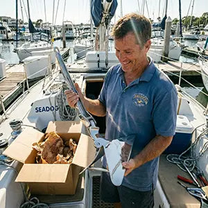 A boat owner on his sailboat deck unboxing an AirowMarine mirror-polished stainless steel anchor, featuring eco-friendly honeycomb paper packaging instead of plastic.